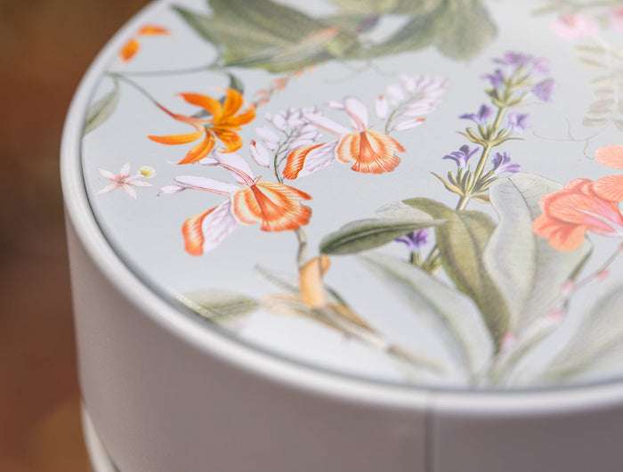 Close-up van een ronde, lichtgekleurde container met een deksel versierd met gedetailleerde, kleurrijke bloemenillustraties, waaronder oranje, roze en paarse bloemen met groene bladeren.
