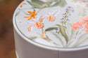Close-up van een ronde, lichtgekleurde container met een deksel versierd met gedetailleerde, kleurrijke bloemenillustraties, waaronder oranje, roze en paarse bloemen met groene bladeren.
