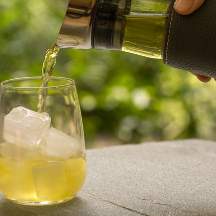 Een hand schenkt een lichtgele drank uit een glazen kan in een helder glas met ijsblokjes, dat buiten op een stenen oppervlak staat, met groen op de achtergrond.