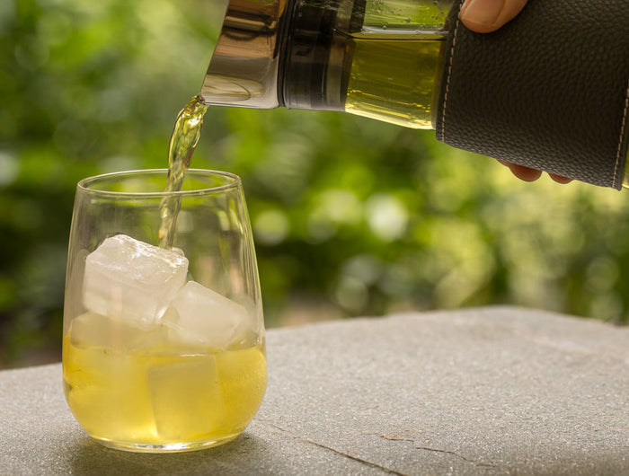 Een hand schenkt een lichtgele drank uit een glazen kan in een helder glas met ijsblokjes, dat buiten op een stenen oppervlak staat, met groen op de achtergrond.