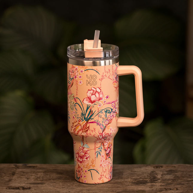 Perzikkleurige geïsoleerde theebeker van 1200 ml met handvat en rietje staat op hout, groene bladeren vaag op de achtergrond.