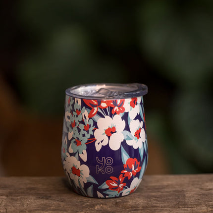 Een dubbelwandige theemok van roestvrijstaal met bloemenmotief en het woord Yoko, op houten ondergrond met groene waas.