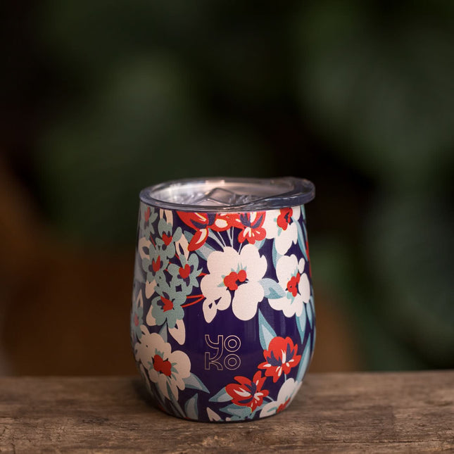 Een dubbelwandige theemok van roestvrijstaal met bloemenmotief en het woord Yoko, op houten ondergrond met groene waas.