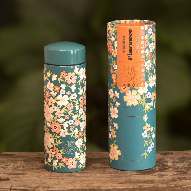 Een turquoise geïsoleerde drinkfles met oranje en crèmekleurig bloemenpatroon staat naast de bijpassende doos op hout, met groene achtergrond.