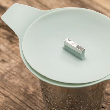 HandyDandy theefilter van metaal met lichtgrijs siliconen deksel en handvat ligt op houten ondergrond, geschikt voor losse thee.