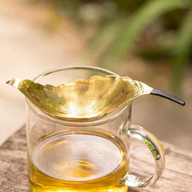 Een roestvrijstalen bladvorm theezeef met zwart handvat ligt op een glas lichte losse thee, op houten ondergrond, met groene planten achteraan.