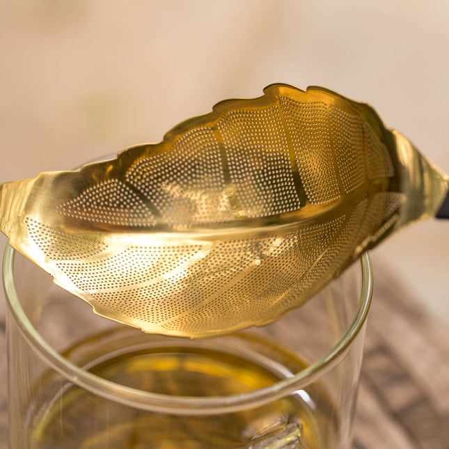 Een roestvrijstalen theezeef in de vorm van een blad ligt op een glas thee, met daarachter een houten oppervlak.