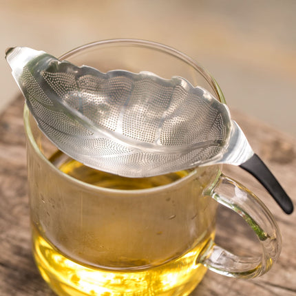 Een bladvormige theezeef van 01Living Teawares roest op een glas met gele losse thee, neergezet op een houten ondergrond.