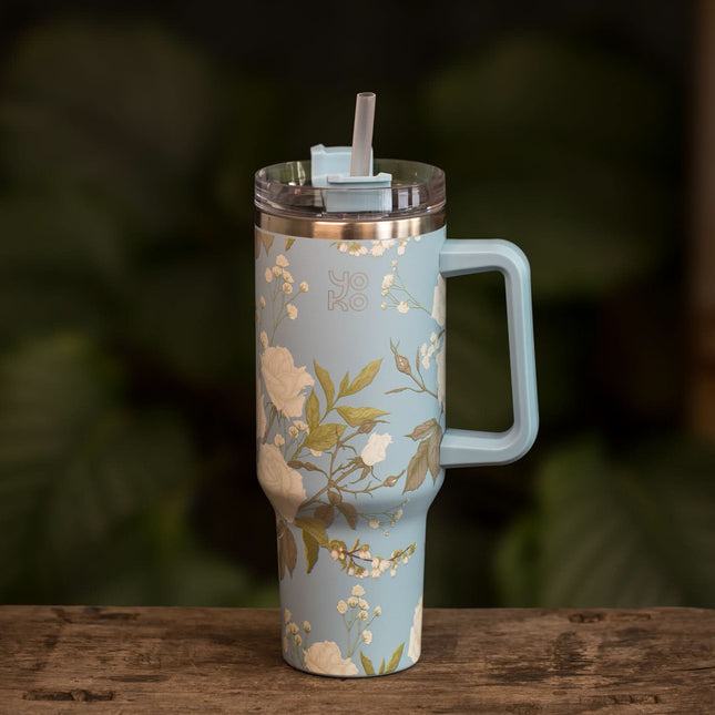 Een lichtblauwe Yoko Design XL thermosbeker met bloemenprint, handvat, deksel en rietje staat op een houten ondergrond.