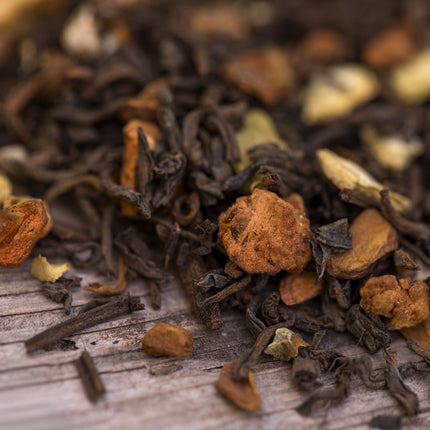 Close-up van De Theeplantage Notenkraker Noire biologisch: losse donkere theebladeren met stukjes noot, gember, kaneel en kardemom op hout.