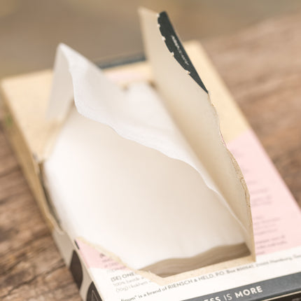Een open, beige met roze tissuebox met een wit tissue staat op een houten ondergrond, naast een Finum-theefilterhouder met 60 filters.