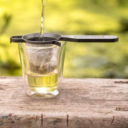Een glas met een theezakje op een houten tafel buiten, waar heet water via een Finum-theefilterhouder met composteerbaar filter wordt ingeschonken.