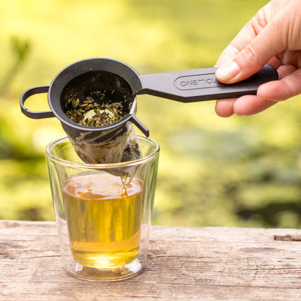 Een hand houdt een Finum-theefilterhouder met composteerbare filters en losse thee boven een glas verse thee op een houten ondergrond, tegen een groene achtergrond.