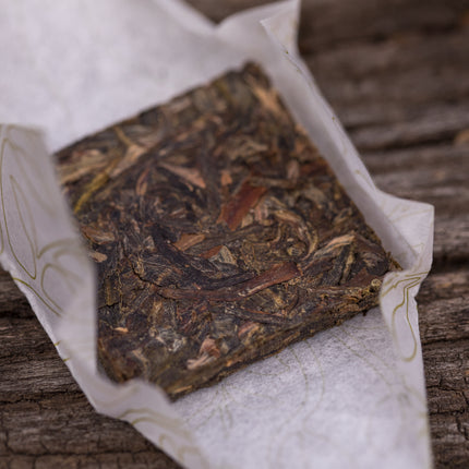 Een Pu-erh Sheng Bio Wu Liang Origin 2021 mini theeblokje, deels in decoratief papier gewikkeld, ligt op een houten ondergrond.