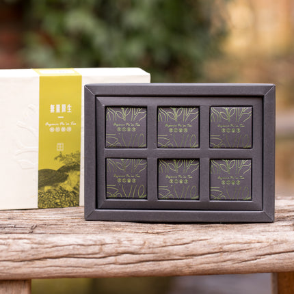 Een doos met acht apart verpakte biologische Pu-erh Sheng Wu Liang mini theecakes in beige en groene verpakking op hout.
