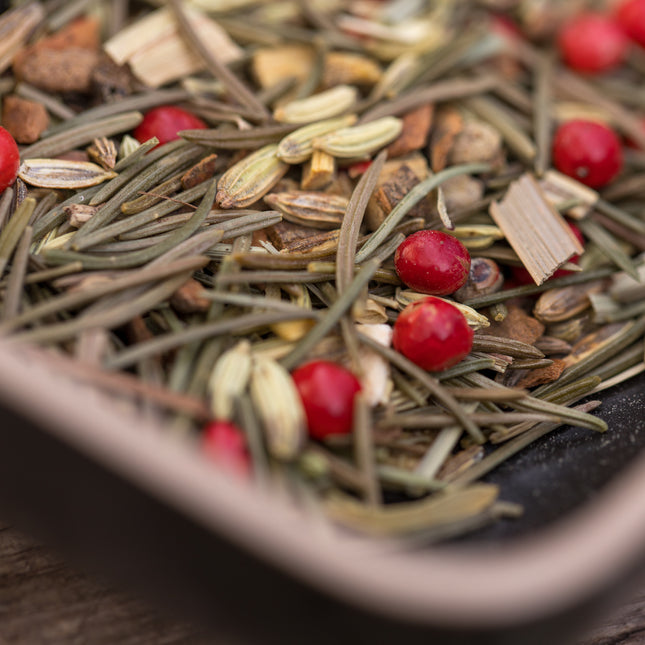 Close-up van losse biologische kruiden voor kerstthee, met groene naaldvormige blaadjes, rode peperkorrels, zaden en stukjes schors in een rechthoekig zwart-beige schaaltje.