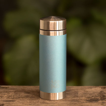 Een lichtblauwe Yoko Design geïsoleerde drinkfles van 350 ml met zilverkleurige dop en bodem staat voor groene bladeren.