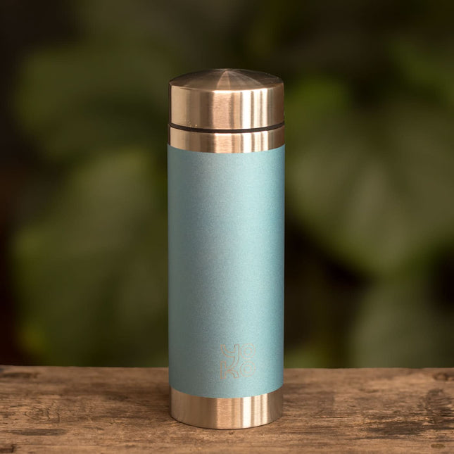 Een lichtblauwe Yoko Design geïsoleerde drinkfles van 350 ml met zilverkleurige dop en bodem staat voor groene bladeren.