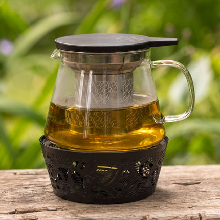 Glazen theepot met infuser en theelichthouder gevuld met thee staat op een houten ondergrond buiten, groene planten op de achtergrond.