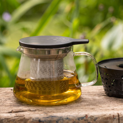 Een glazen theepot met infuser en een theelichthouder staan buiten op hout, gevuld met losse thee; groene planten vervagen op de achtergrond.
