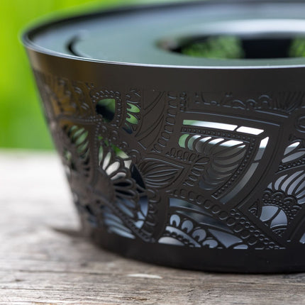 Zwarte metalen theelichthouder met fijn uitgesneden patroon staat op een houten tafel, groene achtergrond is onscherp.
