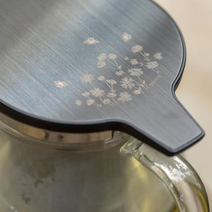 Close-up van een glazen theepot van borosilicaatglas met metalen deksel met gegraveerde bloemen, gevuld met lichte thee.
