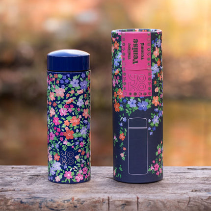 Een geïsoleerde drinkfles met bloemenpatroon en bijpassende verpakking staan op hout, met een vage natuurtuin.