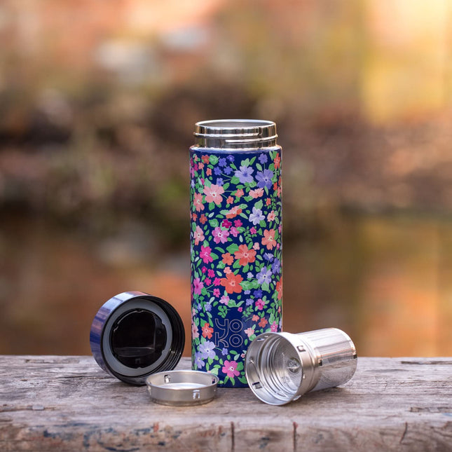 Een geïsoleerde drinkfles van 350 ml met bloemenmotief staat buiten op hout, met deksel, zeef en infuser apart erbij.
