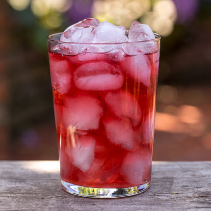 Een helder glas met biologische Berry Easy cold brew thee van De Theeplantage en ijsblokjes staat op een houten ondergrond, buiten vaag in beeld.