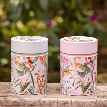 Twee theeblikken van 500 gram met bloemenpatroon, op hout, één met wit en één met roze deksel, groene tuin op de achtergrond.