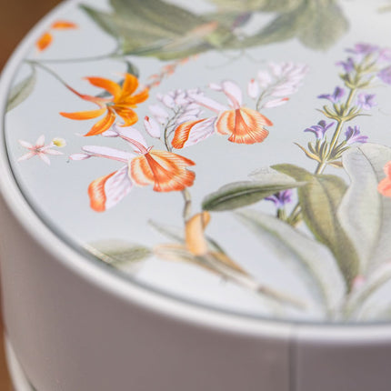 Twee ronde, lichtgekleurde theeblikken van 500 gram met op het deksel bloemen in oranje, roze en paars en groene bladeren.