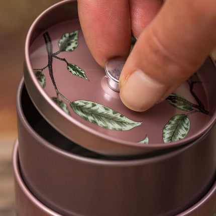 Close-up van een hand die het deksel optilt van een ronde mauve Eigenart theeblik met reliëf van vogels en bladeren.