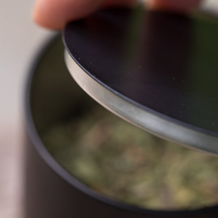 Een hand tilt het zwarte deksel van het ronde bewaarblik van 150 g op, geschikt voor losse thee of kruiden.