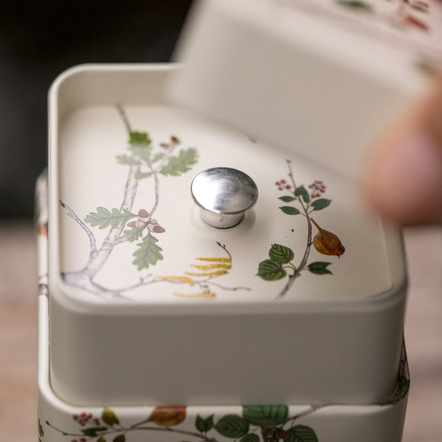 Een hand tilt het deksel op van een van de twee decoratieve Eigenart-theeblikken van 150 gram met een botanisch motief en een metalen knop, geschikt voor losse thee of kruiden.