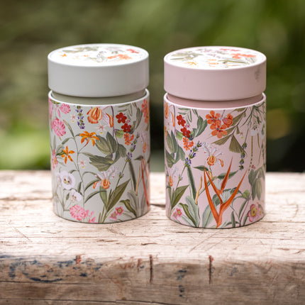 Twee Mira-theeblikken van 150 gram met deksel en botanische bloemenprints staan naast elkaar op een verweerd houten blad met een groene achtergrond.