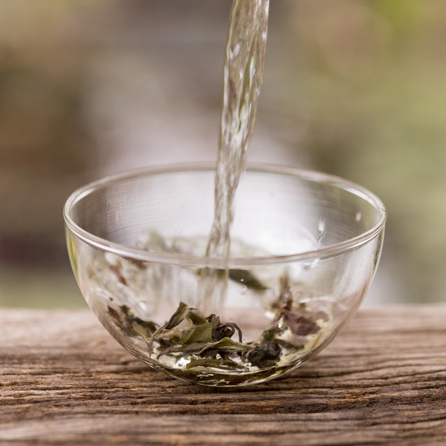 Heet water wordt in glazen theekommen met losse thee geschonken, op een houten ondergrond met zachte natuur op de achtergrond.
