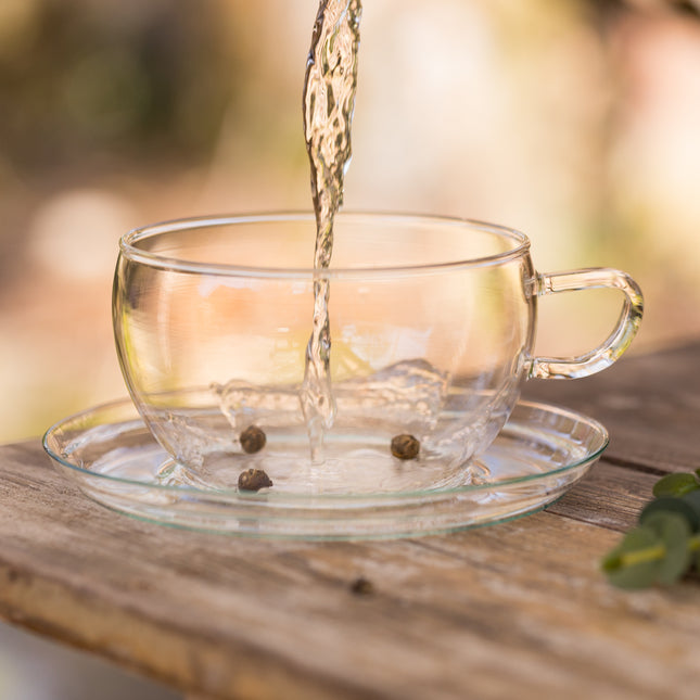 Een heldere vloeistof wordt in een Trendglas Jena theekop met jasmijn parels geschonken, op een houten tafel buiten.