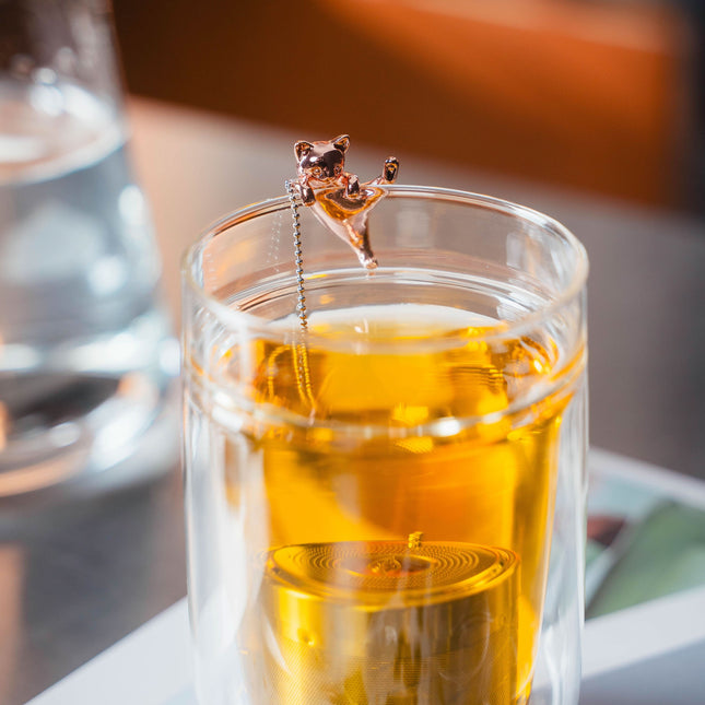 Een glas amberkleurige thee met een metalen katvormig thee-ei hangt aan de rand, naast een glas water op tafel.