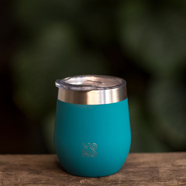 Een geïsoleerde theemok met deksel in blauw van 250 ml staat op hout, met op het mok Yoko Design en vage groene achtergrond.