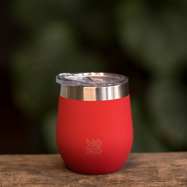 Een rode geïsoleerde theemok van Yoko Design met deksel staat op een houten tafel; op de achtergrond groene bladeren.