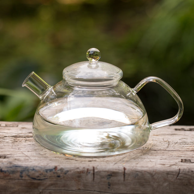Doorzichtige glazen waterkoker van 1,2 liter met rond deksel, buiten op een verweerd houten blad geplaatst, tegen een groene achtergrond; geschikt voor het zetten van thee of kruideninfusies.