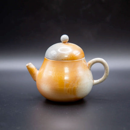 Een ronde, handgemaakte porseleinen theepot met oren en een knopdeksel in oranje-crèmekleur, geplaatst voor een donkere achtergrond.