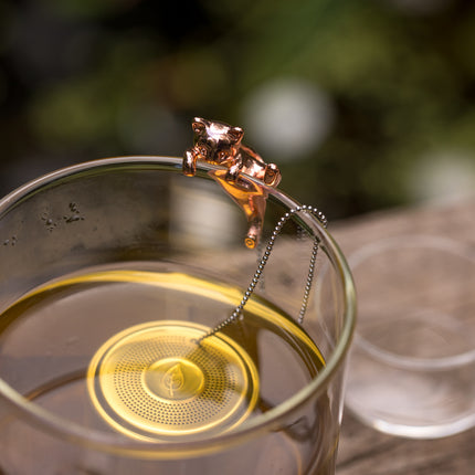 Een roestvrijstalen thee-ei met een kattenfiguurtje hangt gedeeltelijk in een glas losse thee, op de voorgrond tegen een onscherpe achtergrond.