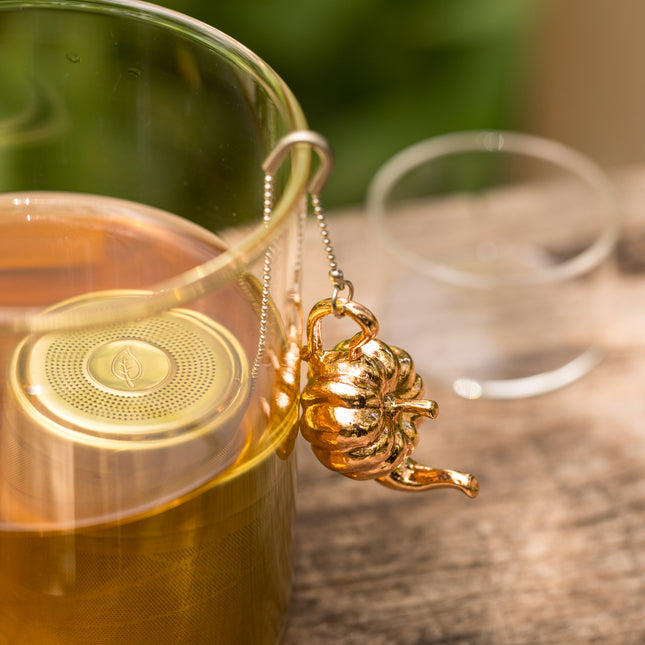 Een glazen theekopje met losse thee staat op een houten ondergrond, met een gouden theeballetje in het kopje en een ketting over de rand.