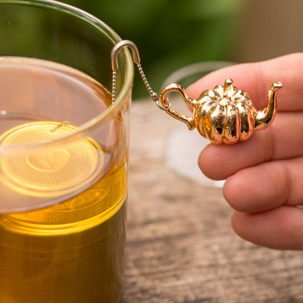Een hand houdt het Pumpkin-thee-ei met glazen lekbakje boven een glas thee; het roestvrijstalen filter hangt in de theekop.