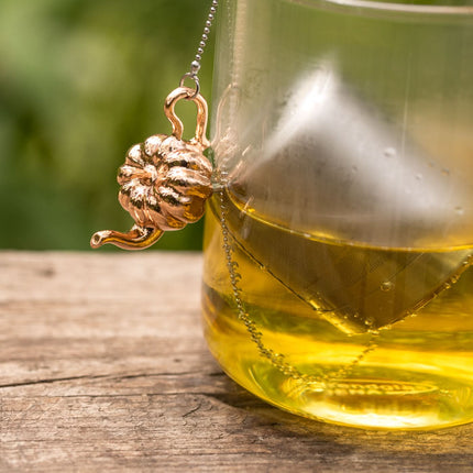 Een glas gele thee met de 01Living Teawares Thee-ei Pumpkin Theepot en glazen lekbakje staan op hout, het kettinkje hangt over de rand.