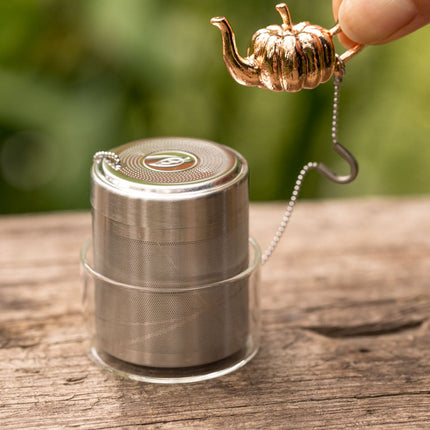 Een hand houdt een metalen thee-ei in pompoenvorm met ketting en glazen lekbakje boven een houten ondergrond met groene achtergrond.