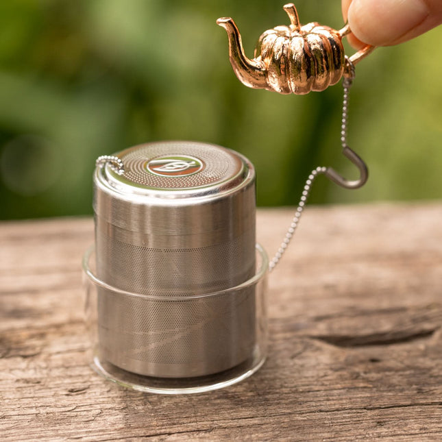 Een hand houdt een metalen thee-ei in pompoenvorm met ketting en glazen lekbakje boven een houten ondergrond met groene achtergrond.