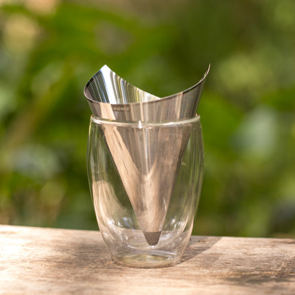 Een RVS theefilter met siliconen rand staat in een dubbelwandig glas op een houten ondergrond, met losse thee en groene achtergrond.