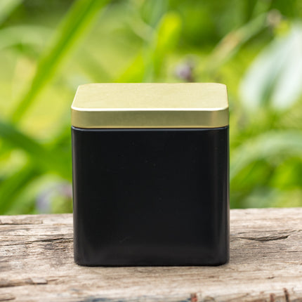 Vierkant zwart-gouden theeblik (9x9x9 cm) van De Theeplantage, afgebeeld tegen een achtergrond van verweerd hout, met groene planten vaag op de achtergrond.